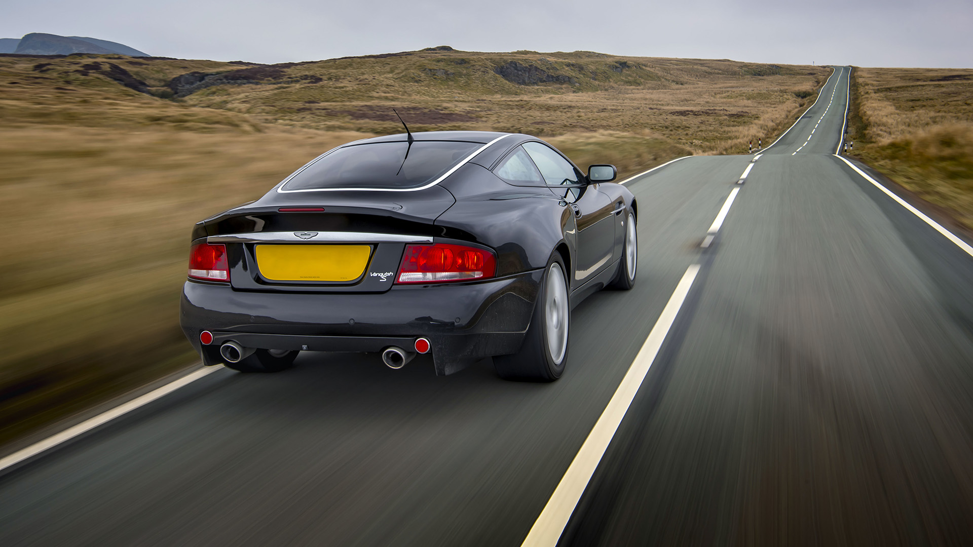 Vista posteriore di un'elegante Aston Martin Vanquish S nera che percorre una strada di campagna panoramica. 