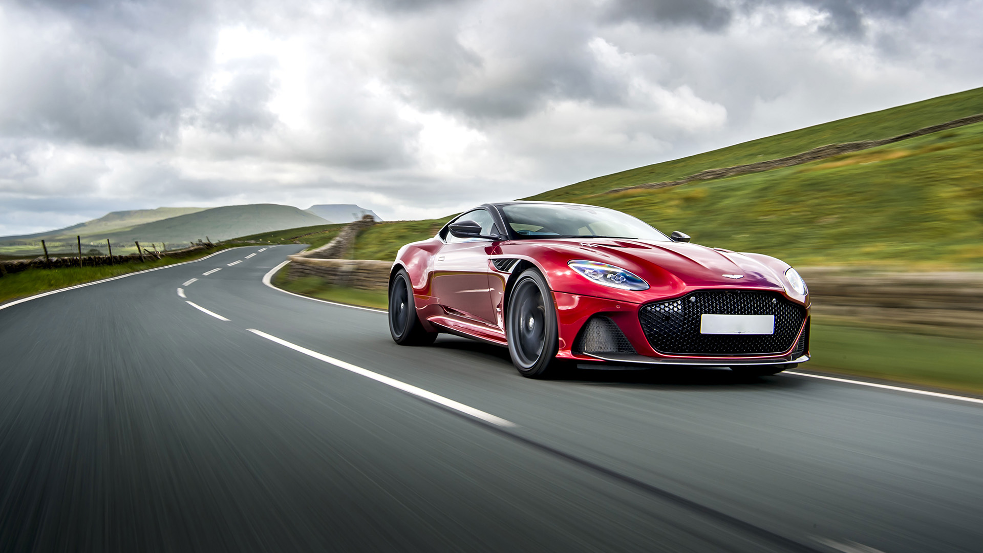 Rosso Aston Martin DBS che guida veloce su una strada di campagna con cielo grigio sopra.