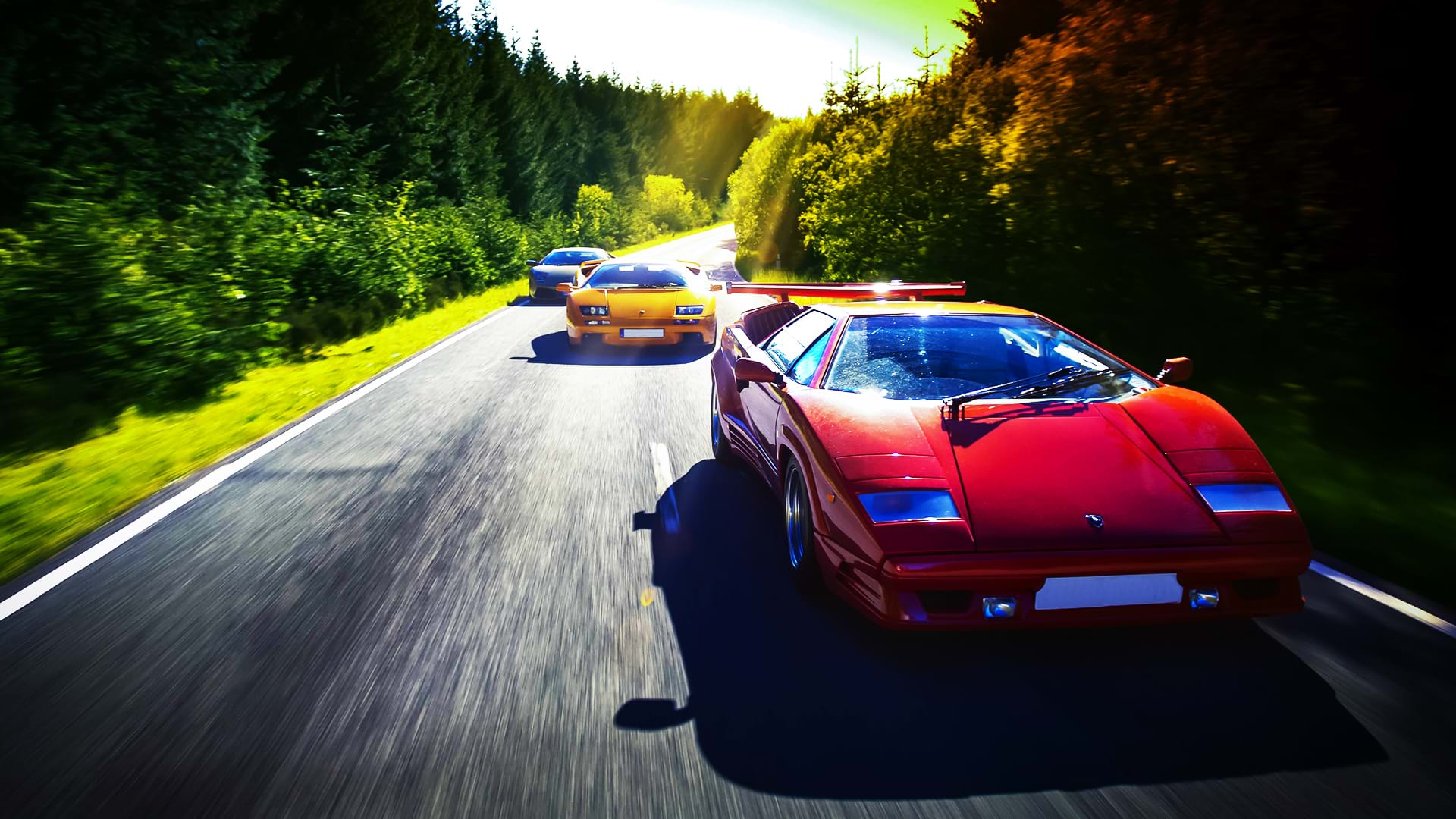 Un colorato convoglio Lamborghini: Countach 25th Anniversary, Diablo 6.0 e Murciélago, corrono lungo una strada panoramica in una giornata luminosa.