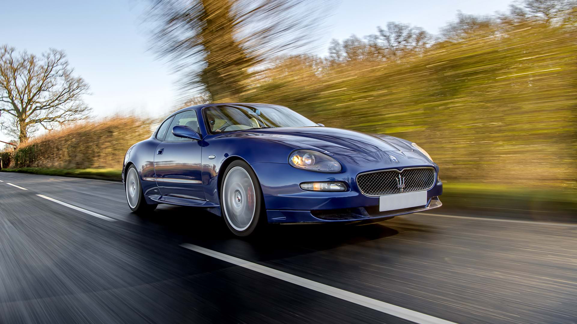 Classic Blue Maserati GranSport che guida lungo una strada di campagna con il cielo azzurro in alto.