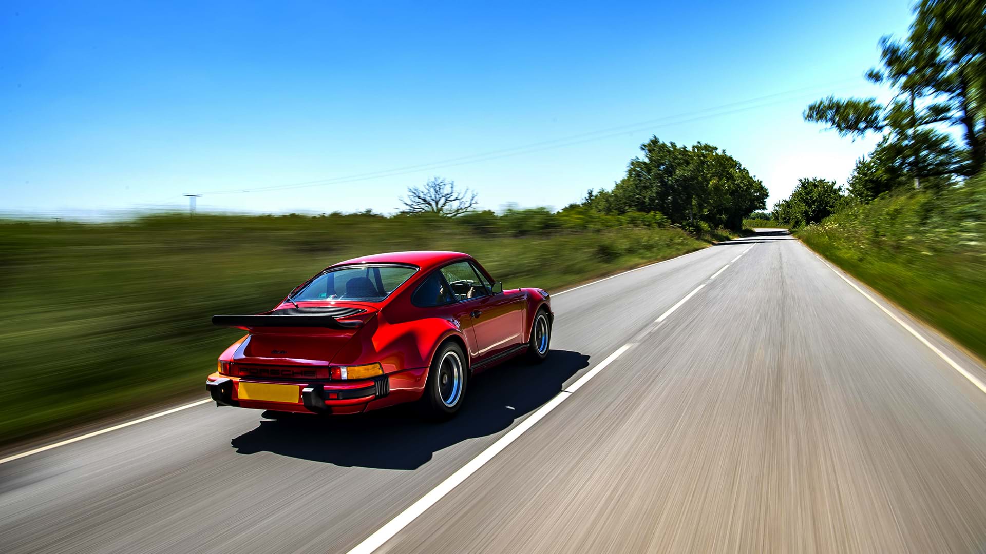 Turbo Porsche 930 rosso vintage che sfreccia lungo una bellissima strada di campagna in una giornata soleggiata.