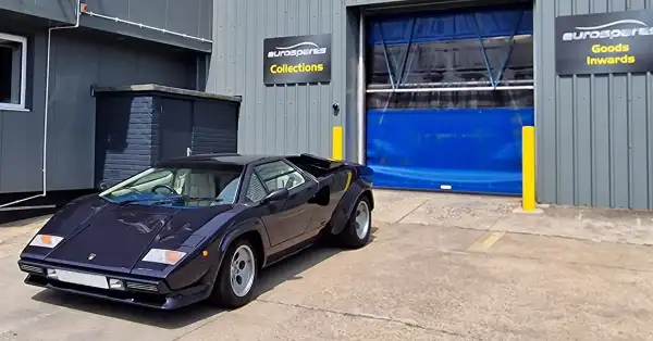 Lamborghini Countach blu parcheggiata fuori dal magazzino Eurospares con cartelli per il ritiro da parte del cliente e per l'accettazione della merce sul muro.